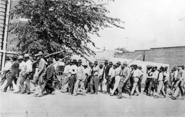 Un grup de trupe Gărzii Naționale, purtând puști cu baioneta atașată, escortează oameni de culoare neînarmați la un centru de detenție după masacrul cursei din Tulsa, Tulsa, Oklahoma, iunie 1921.