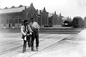Doi militari stau alături de clădirea Pullman și antrenează mașini cu brațele încuiate și o sticlă de lichior în timpul Grevei de Chicago Pullman din 1894