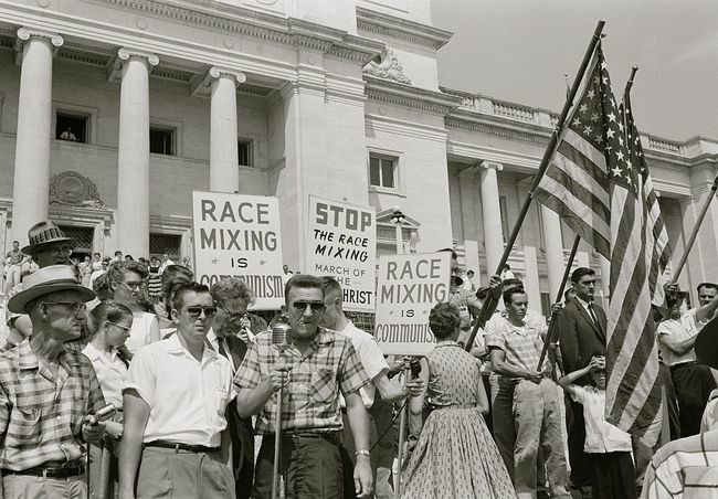 Oameni ținând pancarte și steaguri americane care protestează împotriva admiterii „Little Rock Nine” la Liceul Central.