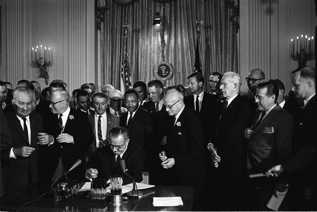 Președintele Lyndon B. Johnson semnează Legea Drepturilor Civile din 1964, în timp ce Martin Luther King, Jr. și alții, urmăresc.