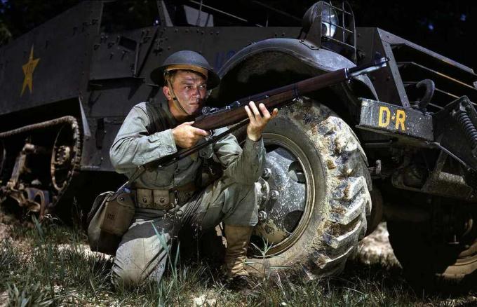 Un soldat american îngenuncheează cu un Garand M1 în fața unui halftrack.